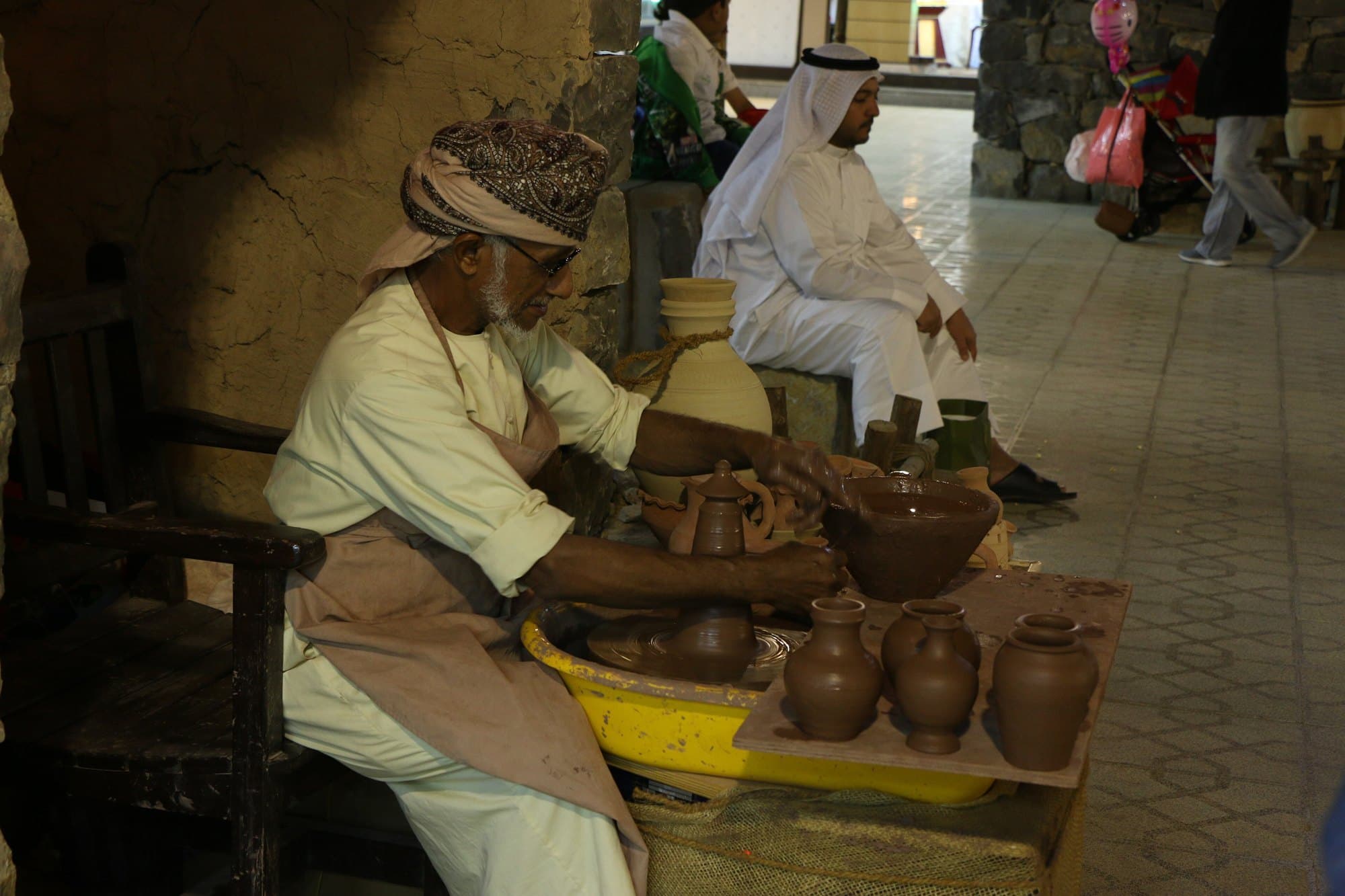 Potter shaping clay on a wheel beside finished pots at Global Village